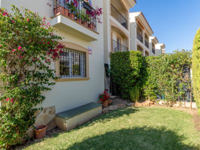 Aperçu du jardin privé avec pelouse verte, bougainvillier fleuri sur la façade et haies. Fenêtre sécurisée et banc extérieur visible.