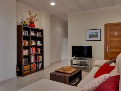Ce salon secondaire polyvalent comprend un canapé confortable, une table basse avec échiquier et une grande bibliothèque garnie de livres. Une télévision et un sol carrelé clair sont visibles.