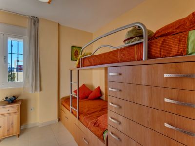 Chambre d'enfants avec lits superposés intégrés dans un meuble en bois clair, incluant des tiroirs de rangement. Une fenêtre avec grille et voilage apporte une lumière naturelle.