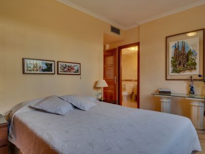 Chambre principale avec lit double, tables de chevet en bois et accès direct à une salle de bain privée. Des œuvres d'art encadrées et une console en verre ornent la pièce.
