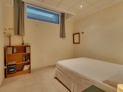 Chambre secondaire avec lit simple, carrelage clair et murs crème. Elle dispose d'une étagère en bois, d'un système audio et d'une fenêtre haute avec rideaux gris.