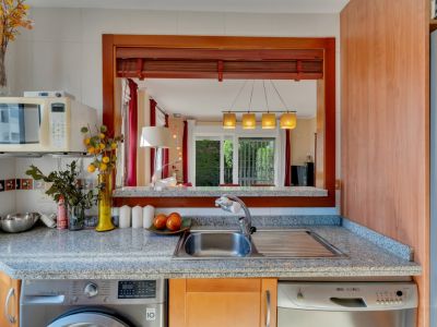 Cuisine fonctionnelle avec plan de travail en granit, évier en acier inoxydable, lave-linge et lave-vaisselle intégrés. Une fenêtre passe-plat en bois s'ouvre sur le salon-salle à manger.