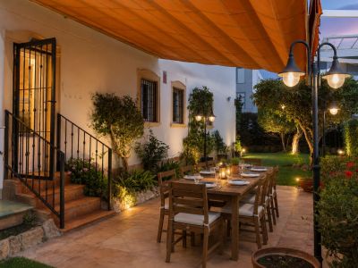 Espace repas extérieur accueillant sur patio carrelé, avec grande table en bois dressée sous un store orange rétractable, entouré d'un jardin illuminé.
