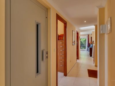 Long couloir aux murs crème et carrelage blanc, doté d'un ascenseur intégré et de plusieurs portes en bois. Au fond, une zone lumineuse avec vue sur le jardin est perceptible.
