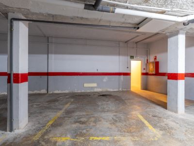 Place de parking souterrain spacieuse avec sol en béton, marquages jaunes et tuyaux apparents. Comprend une porte de sortie éclairée et un équipement anti-incendie.