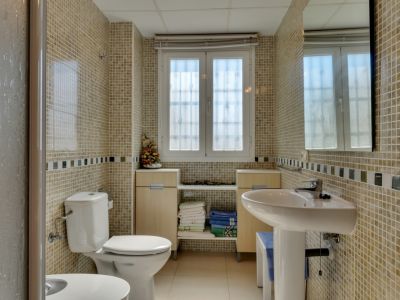 Salle de bain avec murs en mosaïque claire et frise décorative, lavabo sur colonne, toilettes et fenêtre pour la lumière naturelle. Des meubles bas en bois clair offrent du rangement.