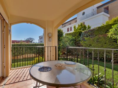 Terrasse couverte avec table en verre et carrelage en terre cuite, offrant une vue directe sur un jardin privé verdoyant avec haies et bâtiments lointains.