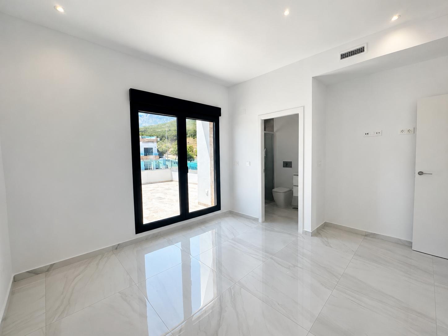 Chambre lumineuse avec carrelage en grès cérame brillant, accès direct à la terrasse et salle de bain attenante. Éclairage encastré et bouche de climatisation visible.