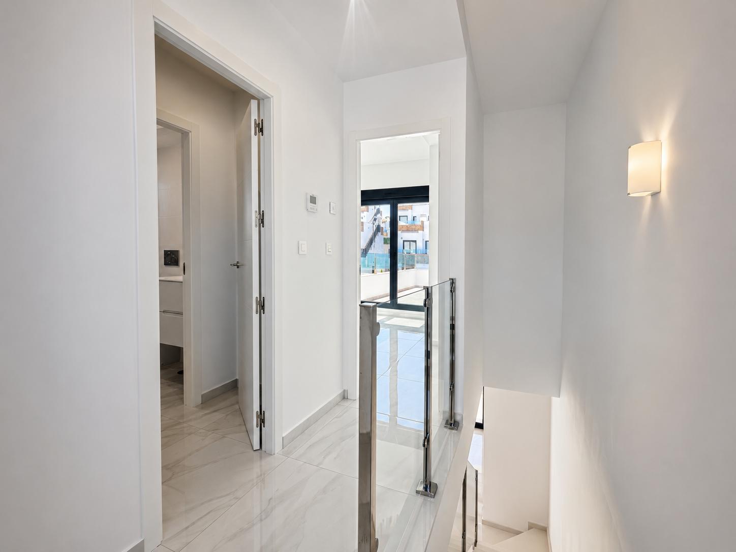 Couloir lumineux à l'étage avec carrelage imitation marbre poli et rampe d'escalier moderne en verre et acier. Une porte mène à une salle de bain, l'autre à un balcon extérieur avec lumière naturelle.