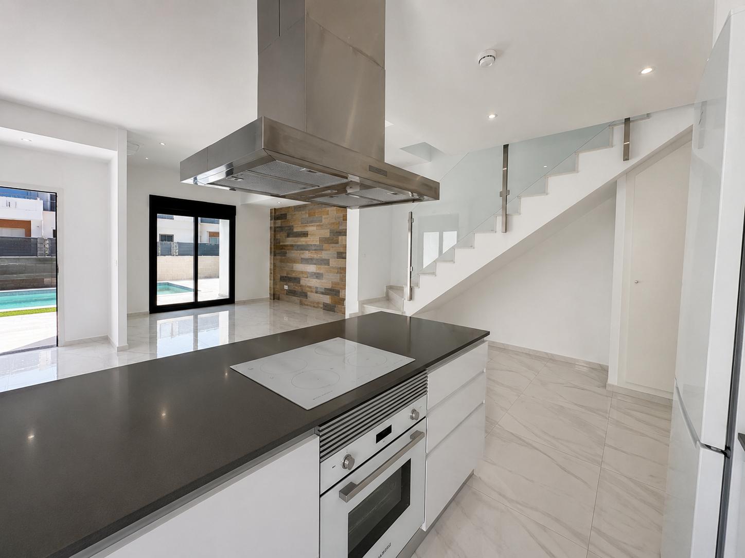 Cuisine ouverte avec îlot à plan de travail foncé, plaque de cuisson intégrée et four. L'espace se connecte au salon et à la piscine, avec un escalier en verre et un mur en pierre.