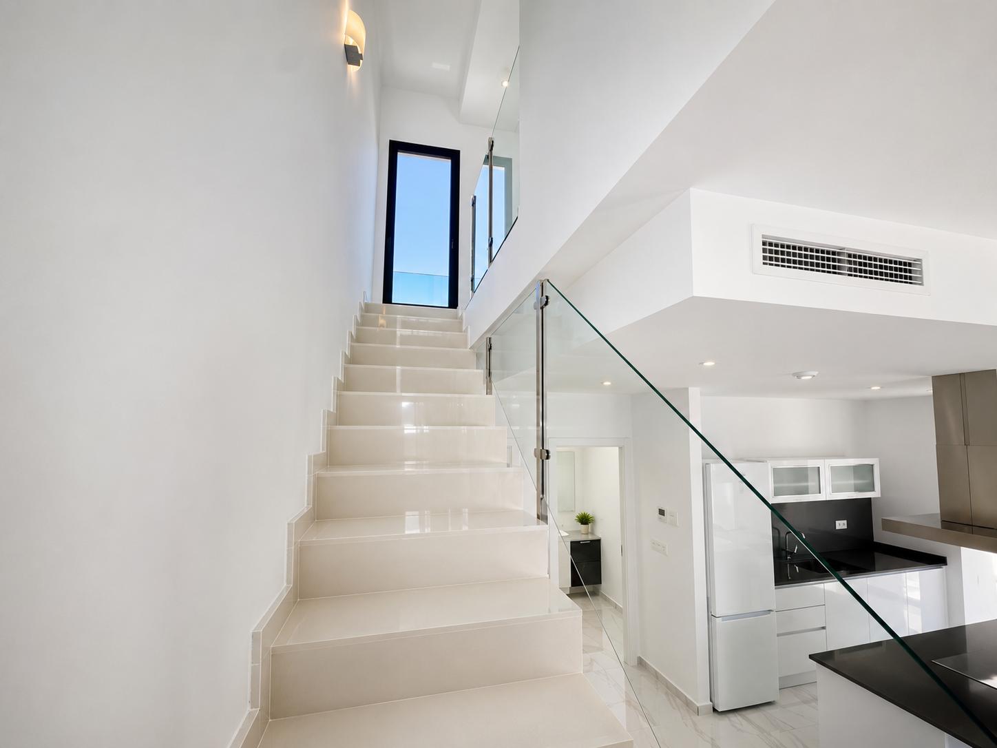 Escalier moderne aux marches claires et rampe en verre élégante, menant à une porte donnant sur l'extérieur. Une cuisine contemporaine et un aperçu d'une salle de bain sont visibles.