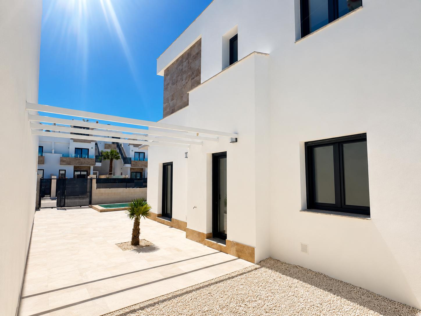 Extérieur de villa moderne avec façade blanche et accents de pierre. Terrasse carrelée claire, petit palmier, pergola blanche et fenêtres à cadre sombre.