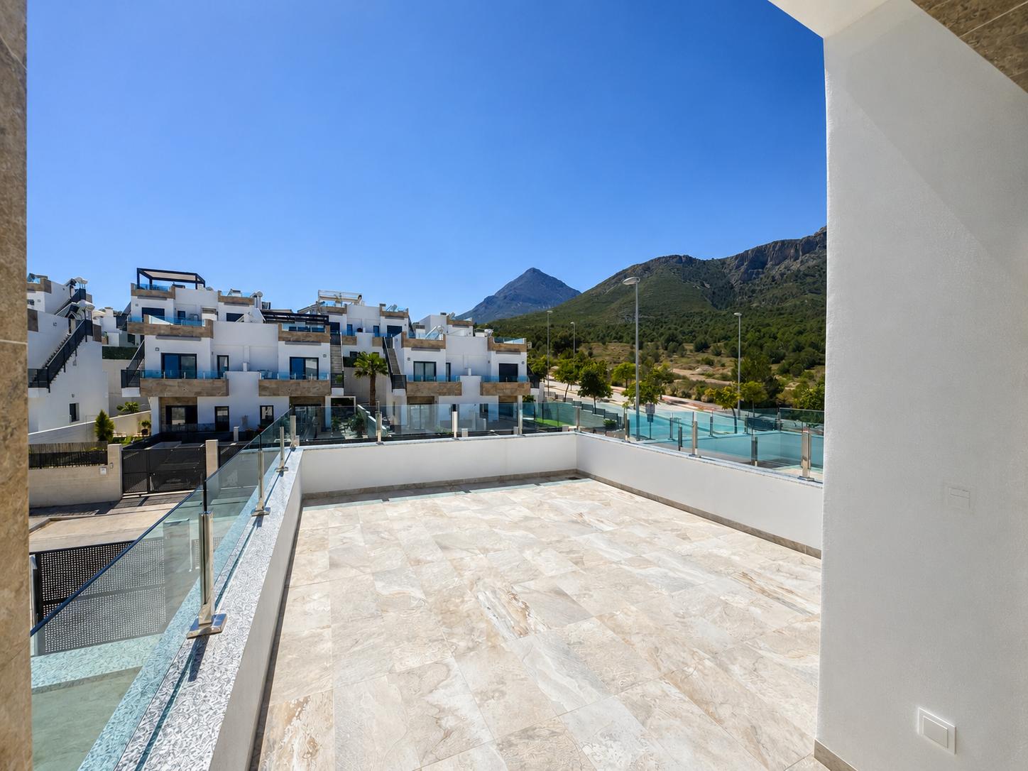 Grande terrasse extérieure avec carrelage clair, garde-corps en verre et vue sur des bâtiments résidentiels modernes et des montagnes boisées sous un ciel bleu dégagé.