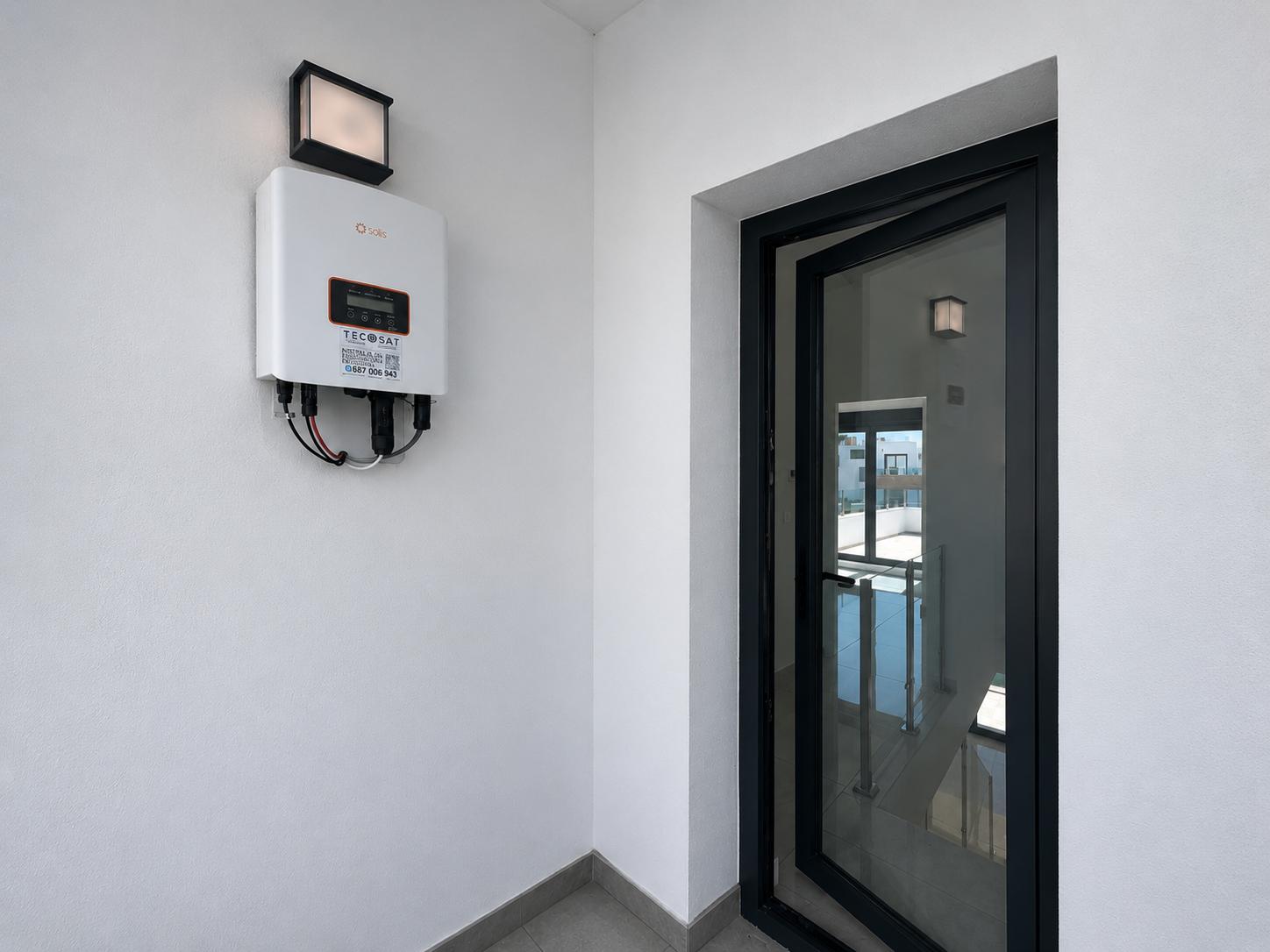 Onduleur solaire Solis fixé sur un mur blanc avec luminaire extérieur. Une porte vitrée à cadre noir entrouverte dévoile un escalier intérieur avec garde-corps.