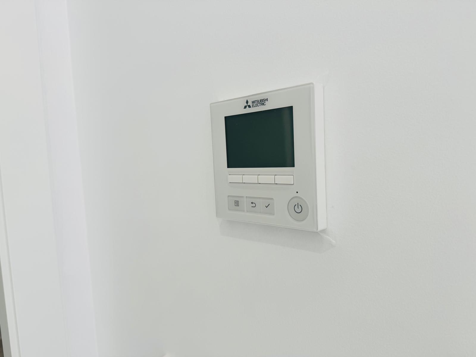 Panneau de commande Mitsubishi Electric pour climatisation gainable, avec écran numérique et boutons intuitifs, intégré au mur blanc.