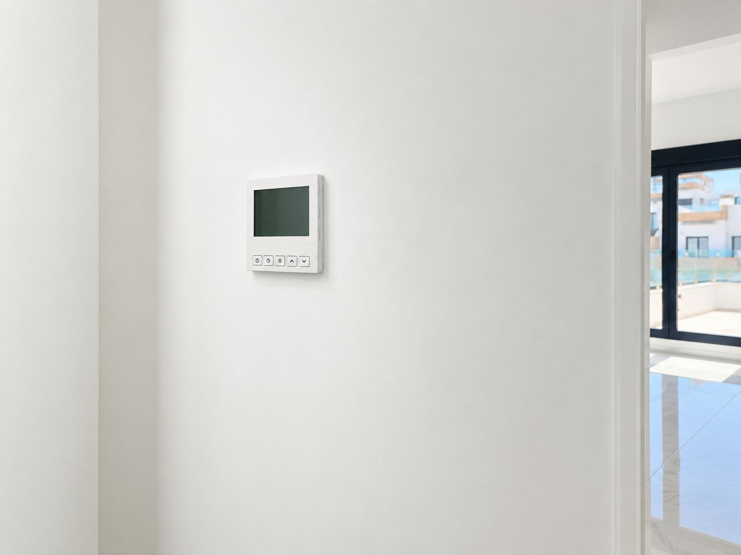Un panneau de commande numérique de climatisation est fixé sur un mur blanc immaculé, offrant un aperçu d'une pièce lumineuse adjacente et d'une terrasse extérieure.