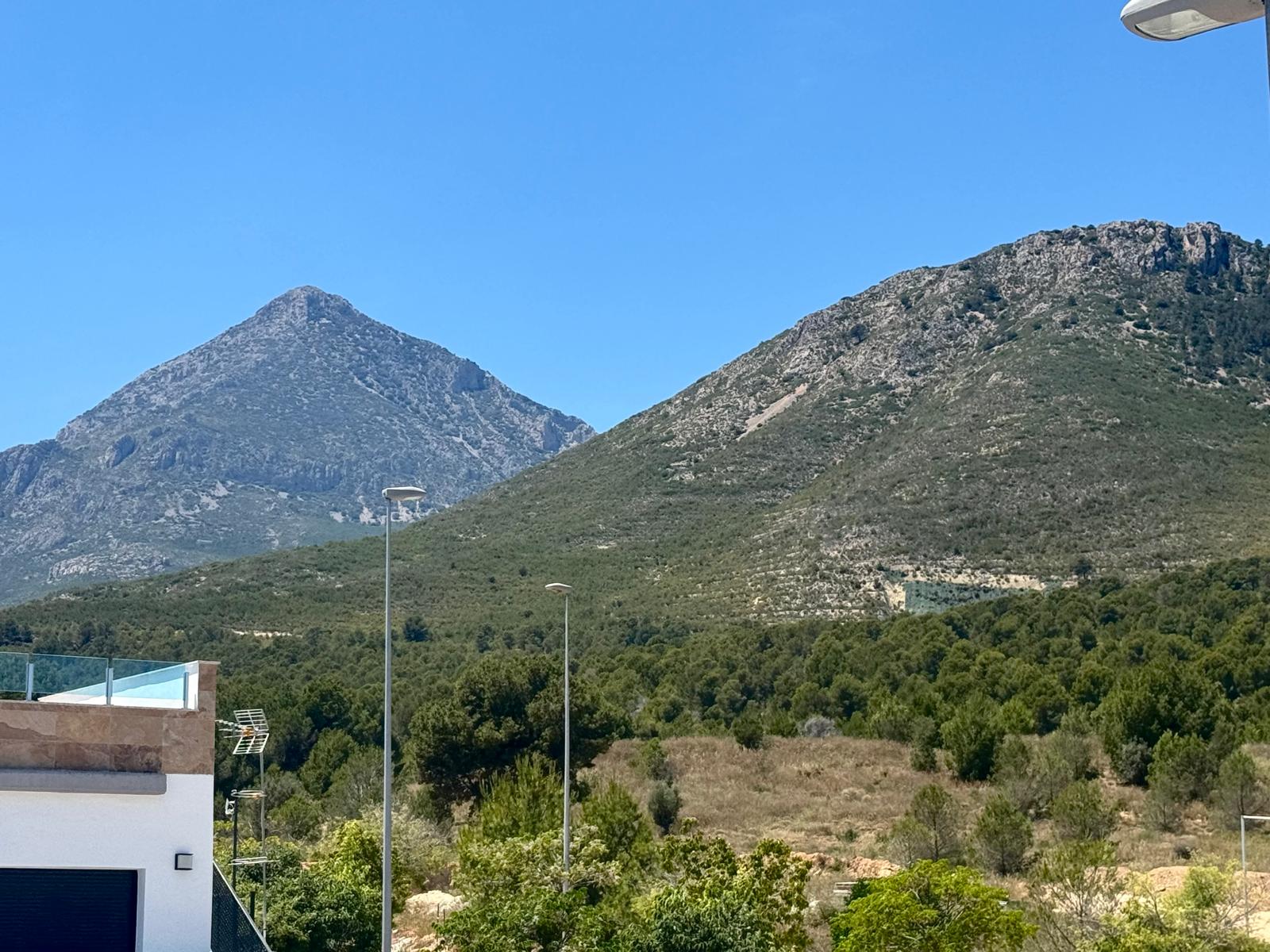 Vues dégagées sur deux montagnes rocheuses distinctes et une végétation méditerranéenne luxuriante, avec une terrasse moderne à balustrade de verre au premier plan.