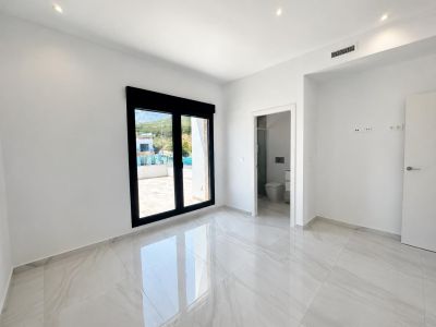 Chambre lumineuse dotée d'un sol carrelé blanc brillant, d'une grande porte coulissante noire ouvrant sur une terrasse et d'un accès à une salle de bain attenante.