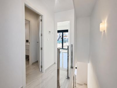 Couloir lumineux à l'étage avec carrelage imitation marbre poli et rampe d'escalier moderne en verre et acier. Une porte mène à une salle de bain, l'autre à un balcon extérieur avec lumière naturelle.