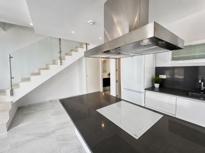 Cuisine ouverte avec îlot central à plan de travail foncé et plaque à induction blanche, jouxtant un escalier moderne avec garde-corps en verre.