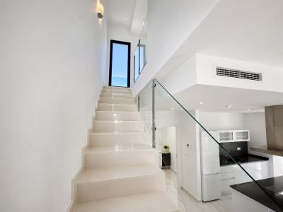 Escalier moderne aux marches claires et rampe en verre élégante, menant à une porte donnant sur l'extérieur. Une cuisine contemporaine et un aperçu d'une salle de bain sont visibles.
