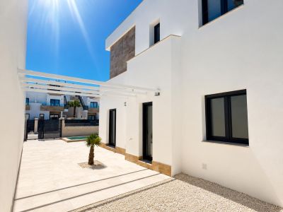Extérieur de villa moderne avec façade blanche et accents de pierre. Terrasse carrelée claire, petit palmier, pergola blanche et fenêtres à cadre sombre.