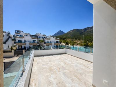 Grande terrasse extérieure avec carrelage clair, garde-corps en verre et vue sur des bâtiments résidentiels modernes et des montagnes boisées sous un ciel bleu dégagé.