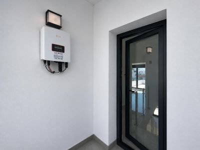 Onduleur solaire Solis fixé sur un mur blanc avec luminaire extérieur. Une porte vitrée à cadre noir entrouverte dévoile un escalier intérieur avec garde-corps.