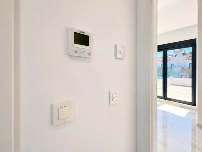 Panneau de contrôle de climatisation Mitsubishi et interrupteurs modernes sur mur blanc, à côté de grandes portes coulissantes menant à une terrasse carrelée avec garde-corps en verre.