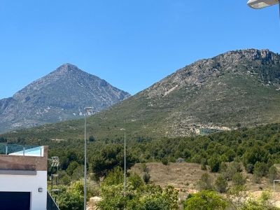 Vues dégagées sur deux montagnes rocheuses distinctes et une végétation méditerranéenne luxuriante, avec une terrasse moderne à balustrade de verre au premier plan.