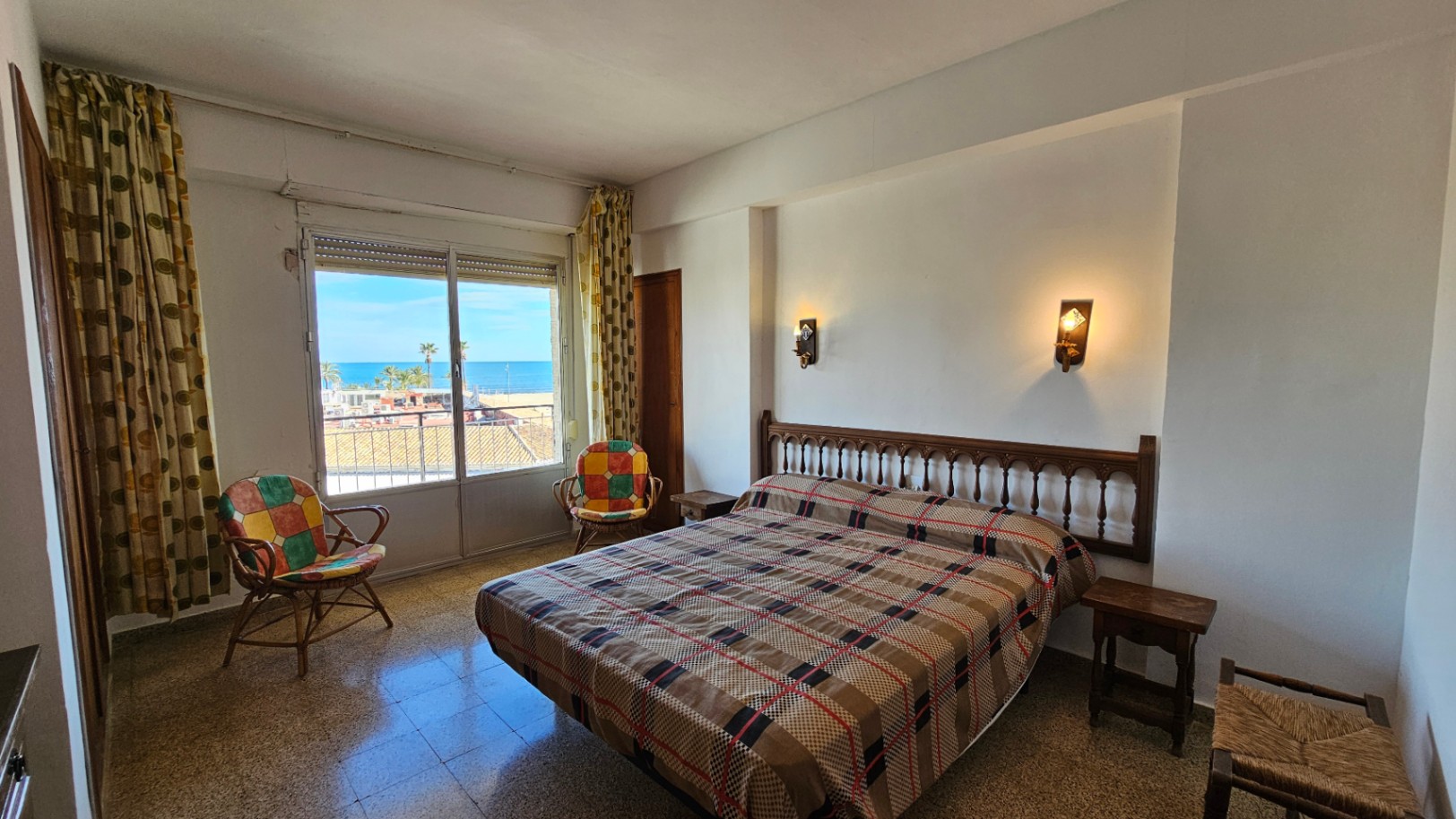 Chambre à coucher avec lit double et tête de lit en bois sculpté, offrant une vue directe sur la mer Méditerranée depuis sa fenêtre avec store et rideaux à motifs. Sol en terrazzo.