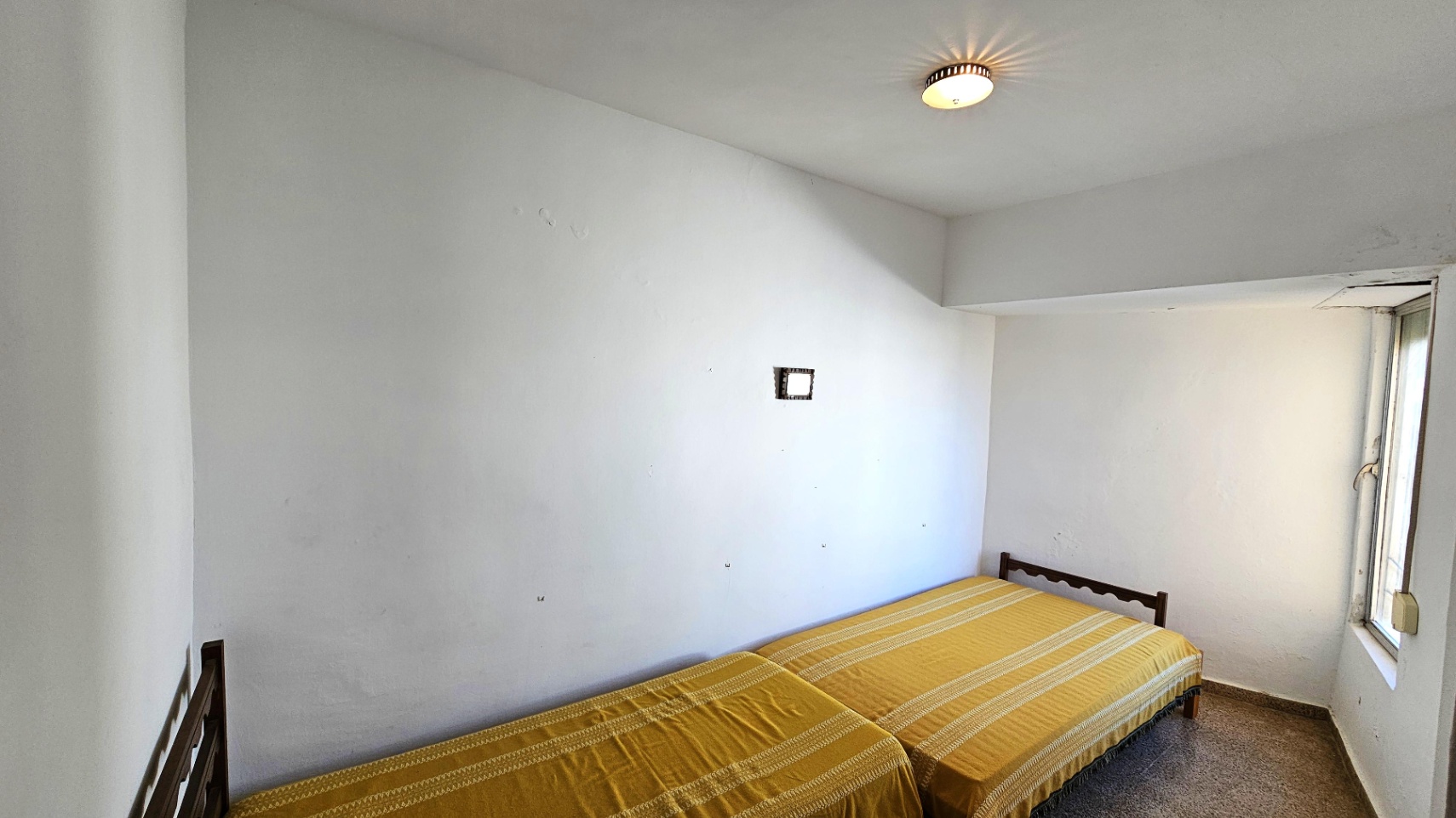 Chambre avec deux lits simples à cadres en bois. Les murs blancs présentent des traces d'usure, éclairée par une fenêtre latérale et un sol en terrazzo.