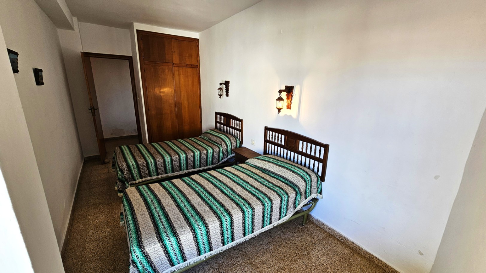 Chambre avec deux lits simples à têtes de lit en bois et couvre-lits rayés. Elle dispose d'une armoire encastrée en bois et d'appliques murales, sur un sol carrelé.
