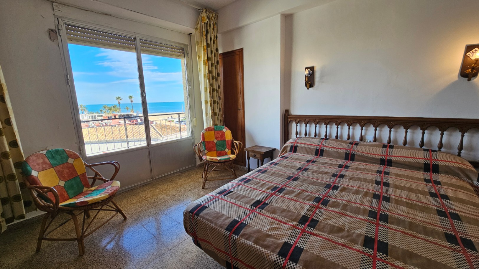 Chambre avec vue mer directe depuis une grande fenêtre, lit avec tête de lit en bois, couvre-lit à carreaux et deux fauteuils en rotin. Lumière naturelle sur le sol carrelé.