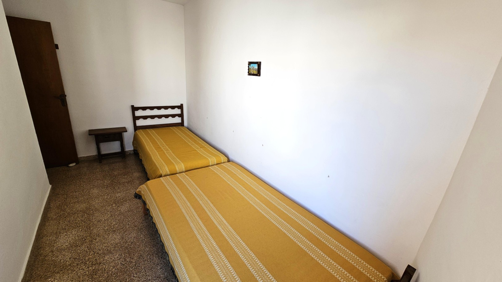 Chambre simple avec deux lits jumeaux, des couvre-lits jaunes à rayures et une tête de lit en bois foncé. Une petite table de chevet et un sol granuleux.