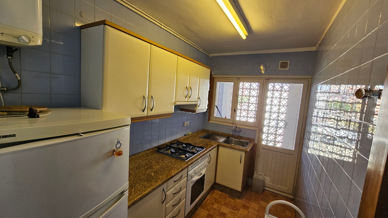 Cuisine fonctionnelle avec carrelage bleu, plan de travail effet granit et meubles blancs. Équipée d'une plaque de cuisson à gaz, d'un four et d'une fenêtre à motifs.