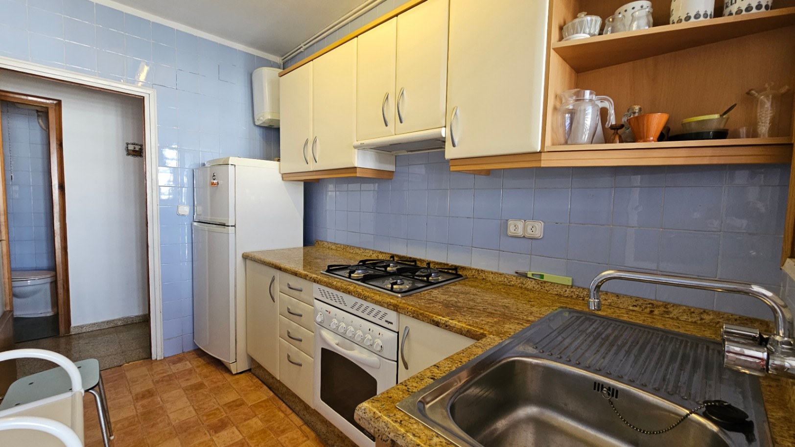 Cuisine fonctionnelle avec carrelage mural bleu clair, plan de travail effet granit et armoires blanches. Équipée d'une plaque de cuisson à gaz, four, évier en inox et réfrigérateur.