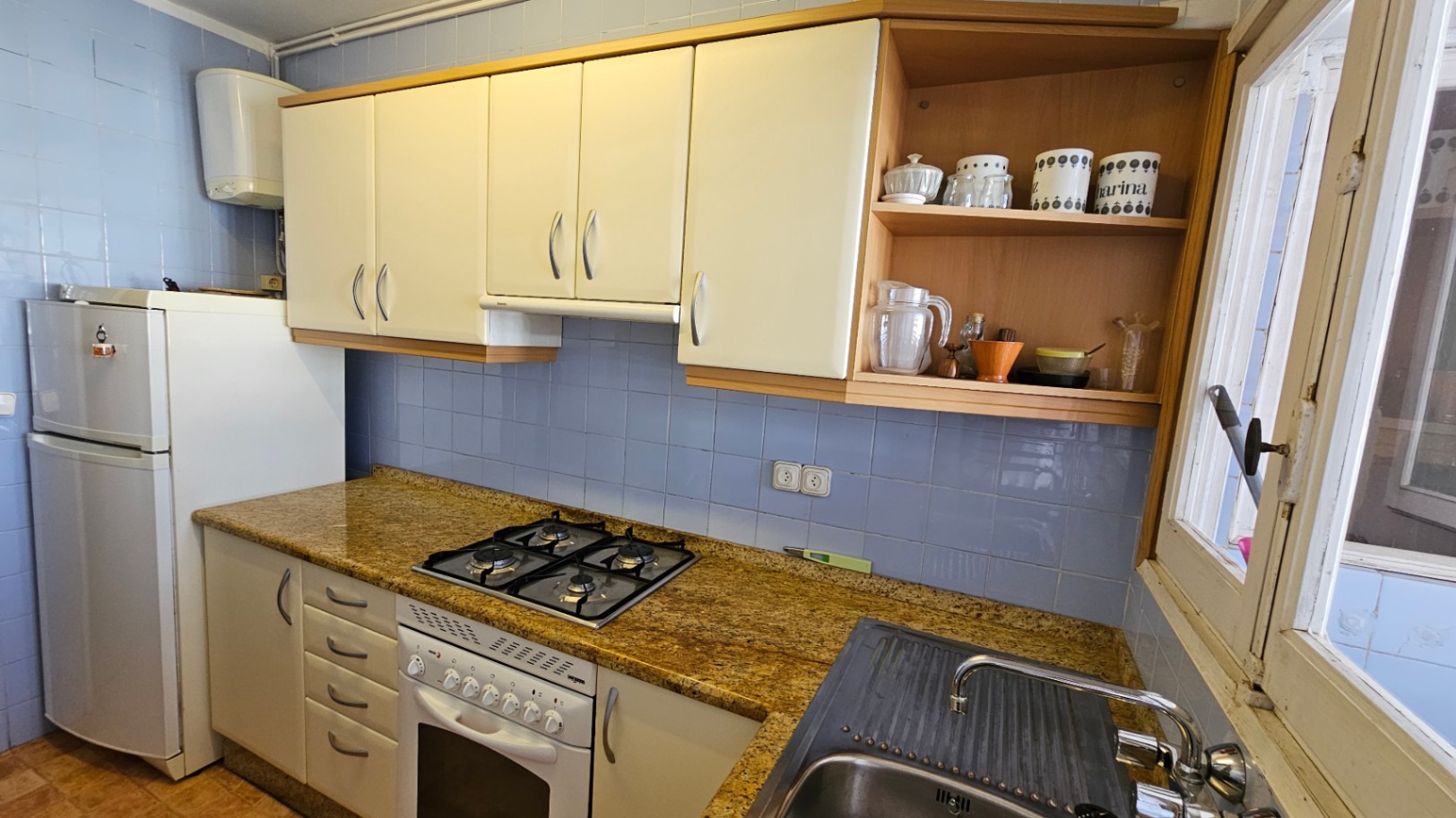 Cuisine fonctionnelle avec carrelage mural bleu, plan de travail en granit moucheté et armoires crème. Équipée d'une plaque de cuisson à gaz, four et évier en inox, éclairée par une fenêtre.