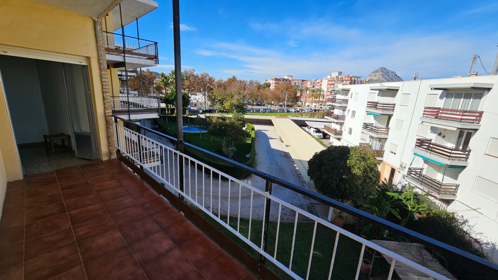 Grand balcon carrelé de terre cuite, avec vue sur le jardin commun, la piscine, les bâtiments résidentiels et une montagne lointaine sous un ciel clair.