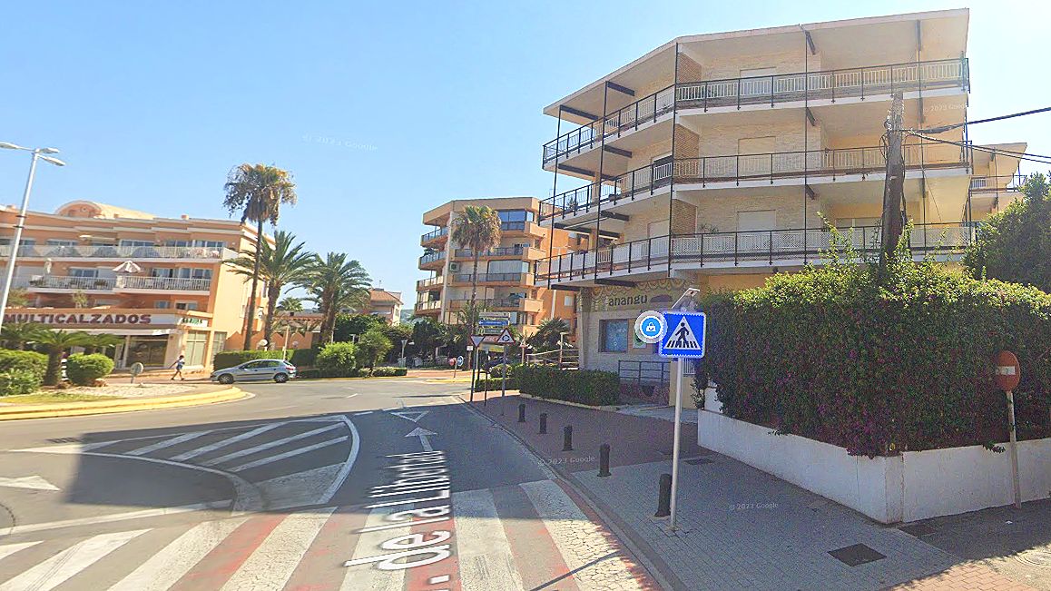 Immeuble résidentiel d'angle avec façade claire et balcons multiples, situé près d'un carrefour avec passage piéton et végétation dense.