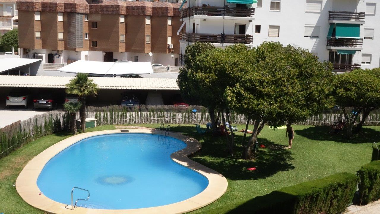 Piscine commune ovale avec margelle claire, entourée d'une pelouse verte et d'arbres ombragés. Un parking couvert et des bâtiments résidentiels sont visibles à l'arrière-plan.