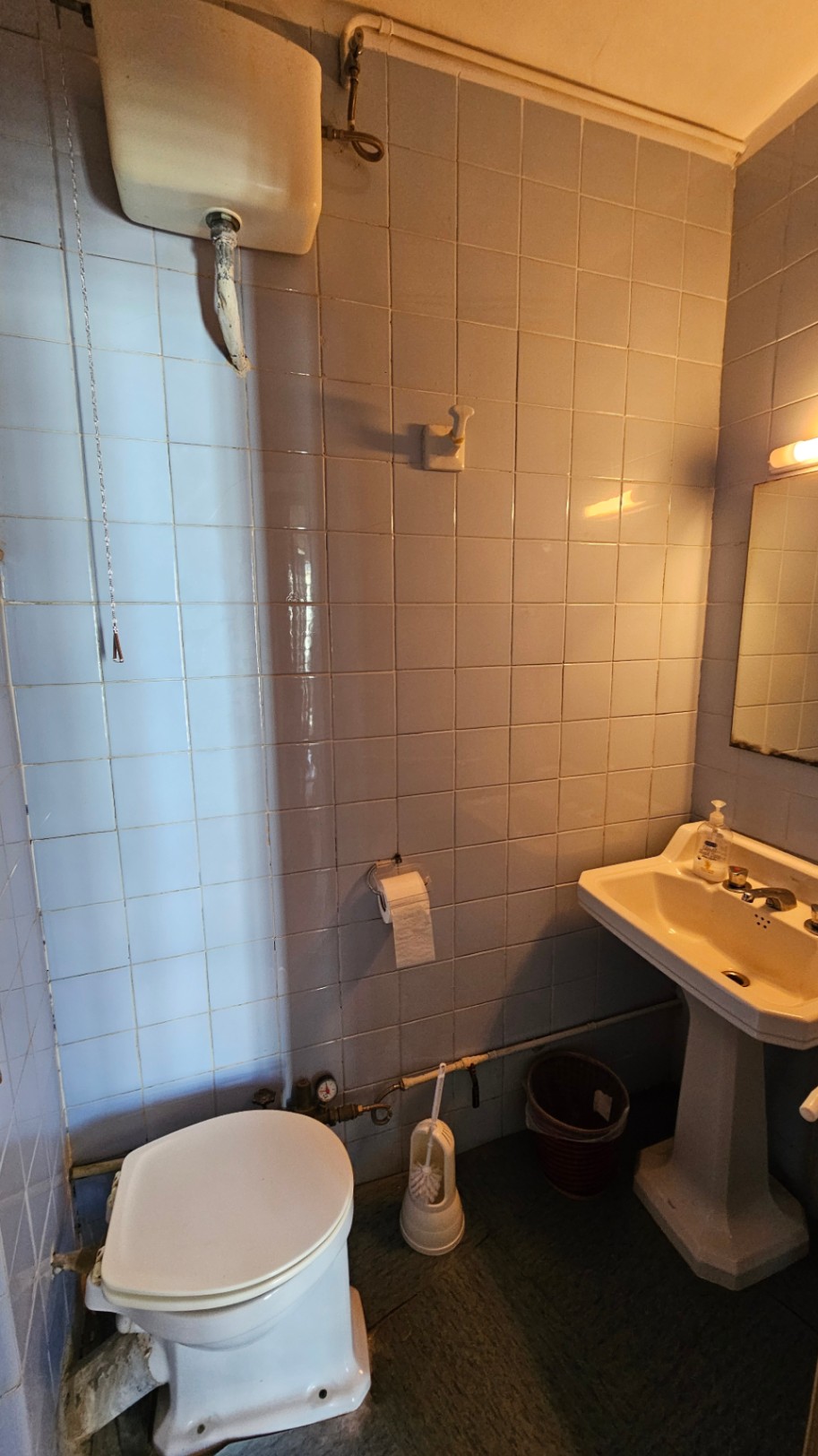 Salle de bain compacte avec carrelage mural carré bleu clair, réservoir de chasse haut à chaîne, lavabo sur colonne et plomberie apparente. Miroir et applique murale.
