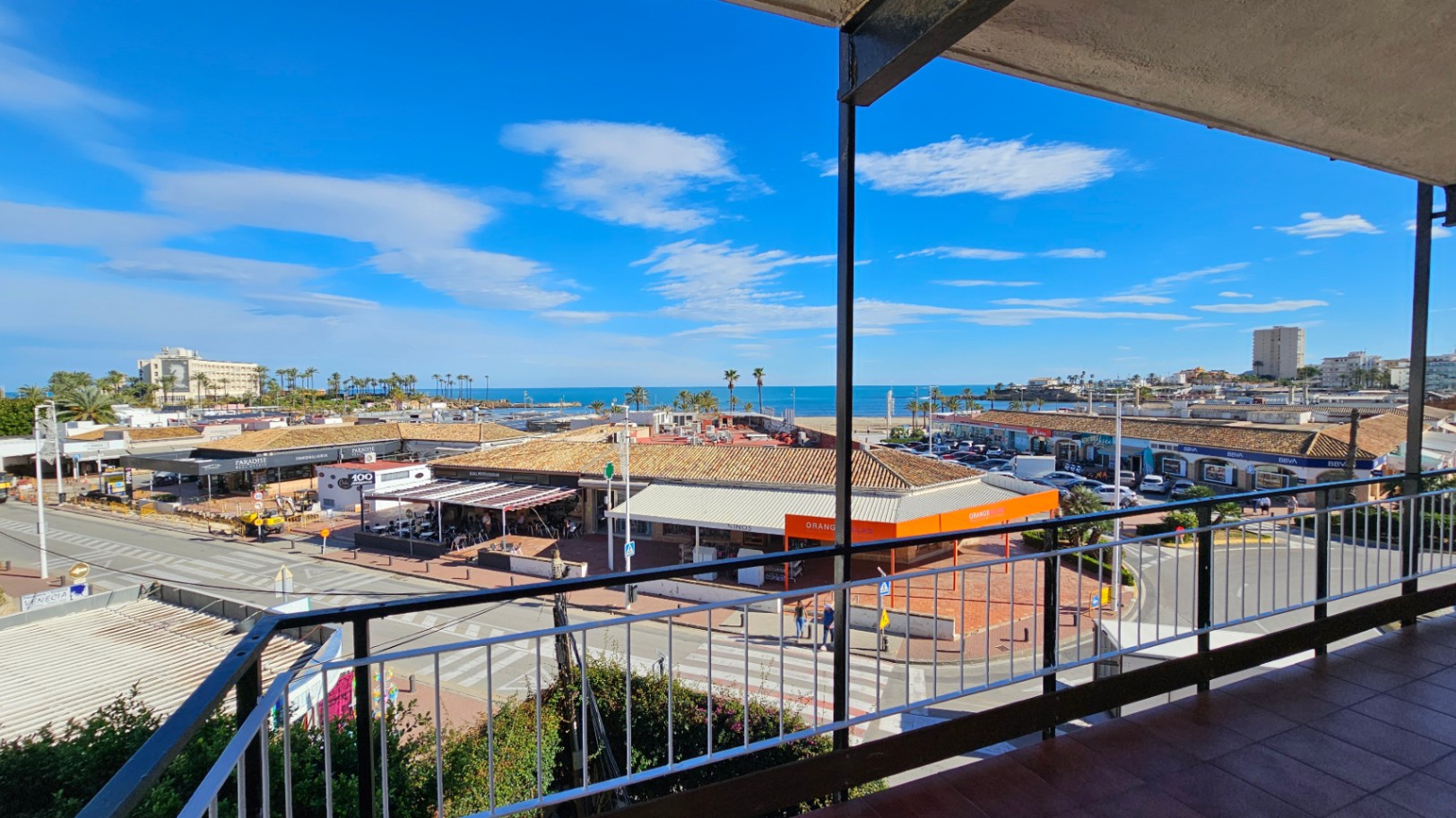 Vue depuis la terrasse sur une rue animée avec des commerces et des toits de tuiles traditionnels, s'étendant vers la plage de l'Arenal et la mer Méditerranée sous un ciel clair.
