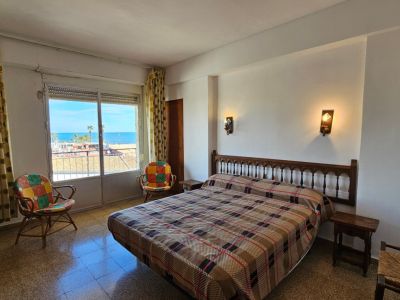 Chambre à coucher avec lit double et tête de lit en bois sculpté, offrant une vue directe sur la mer Méditerranée depuis sa fenêtre avec store et rideaux à motifs. Sol en terrazzo.