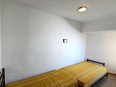 Chambre avec deux lits simples à cadres en bois. Les murs blancs présentent des traces d'usure, éclairée par une fenêtre latérale et un sol en terrazzo.