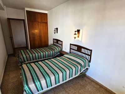 Chambre avec deux lits simples à têtes de lit en bois et couvre-lits rayés. Elle dispose d'une armoire encastrée en bois et d'appliques murales, sur un sol carrelé.