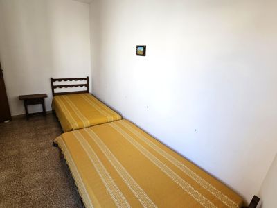 Chambre simple avec deux lits jumeaux, des couvre-lits jaunes à rayures et une tête de lit en bois foncé. Une petite table de chevet et un sol granuleux.
