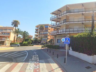 Immeuble résidentiel d'angle avec façade claire et balcons multiples, situé près d'un carrefour avec passage piéton et végétation dense.
