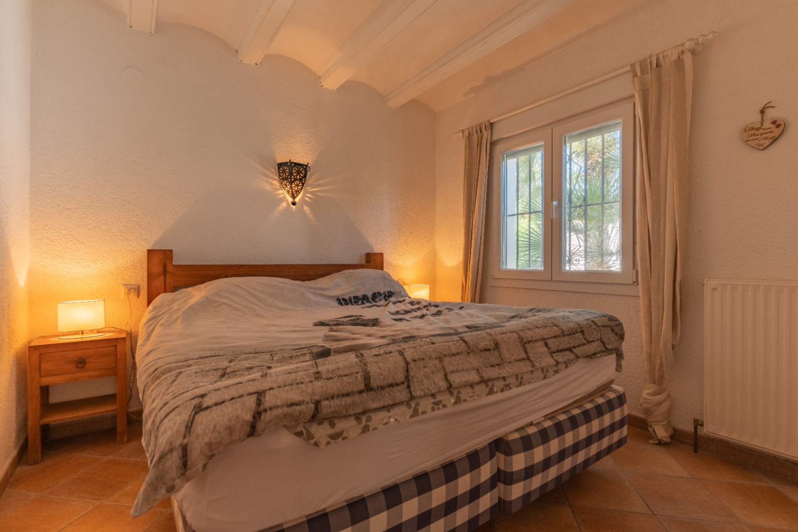 Chambre à coucher avec lit en bois et base à carreaux, sol en terre cuite et fenêtre PVC à double vitrage. Une applique murale décorative projette des motifs lumineux, complétant le plafond à poutres apparentes.