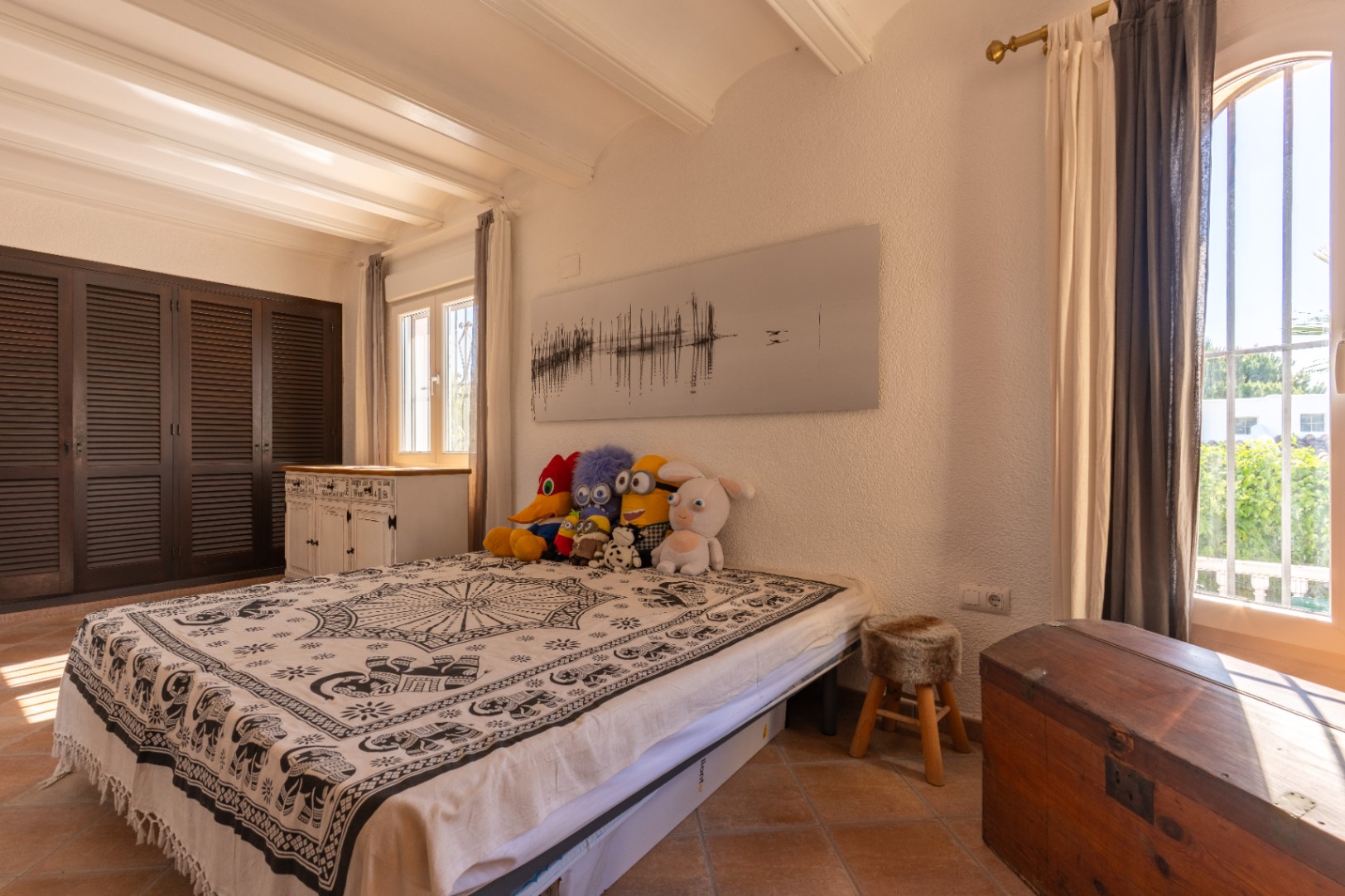 Chambre lumineuse avec sol en terre cuite, murs blancs texturés et poutres apparentes. Armoires à persiennes foncées, lit avec couverture à motifs et lumière naturelle des fenêtres PVC double vitrage.